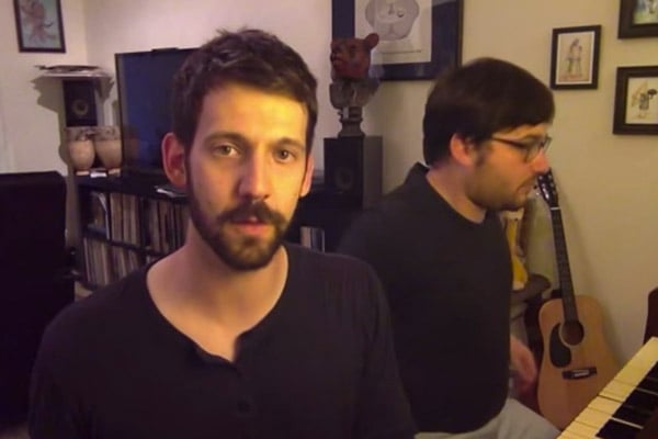 Two perfect men showcasing celebrity impressions next to a piano in a living room.