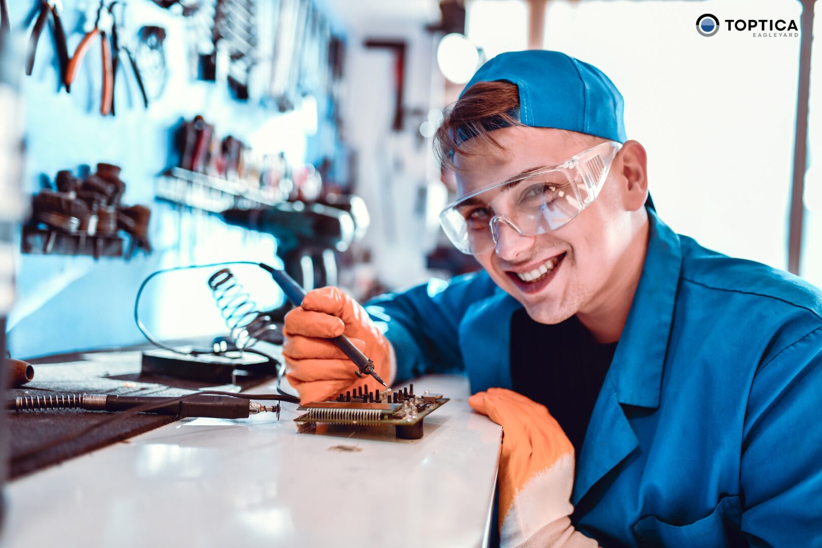 A Werkstudent in blue uniform, safety goggles, and orange gloves works on an electronic circuit board at a workstation with tools in the background.