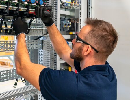 A person wearing gloves and protective glasses, likely an Elektrotechniker, is meticulously working on wiring inside an electrical panel.